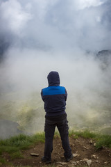 Hiker with blue jacket and hood over head standing at the edge of Lake peak and looking at seven Rila lakes through thick fog