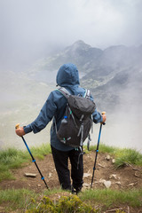 Hiker with blue jacket with hood over head, walking sticks and backpack standing at the edge of Lake peak and looking at seven Rila lakes through thick fog