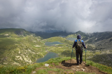 Hiker with blue jacket with hood over head, walking sticks and backpack standing at the edge of Lake peak and looking at seven Rila lakes through thick fog