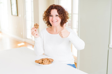 Senior woman eating chocolate chips cookies at home happy with big smile doing ok sign, thumb up with fingers, excellent sign