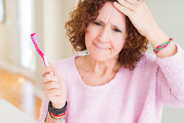 Senior woman holding pink toothbrush at dental clinic stressed with hand on head, shocked with shame and surprise face, angry and frustrated. Fear and upset for mistake.