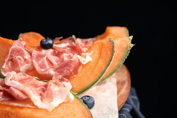Wooden board with melon, prosciutto and blueberries on table against black background