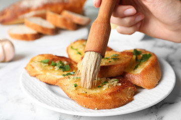 Woman brushing slices of delicious toasted bread with garlic and herbs on white marble table, closeup