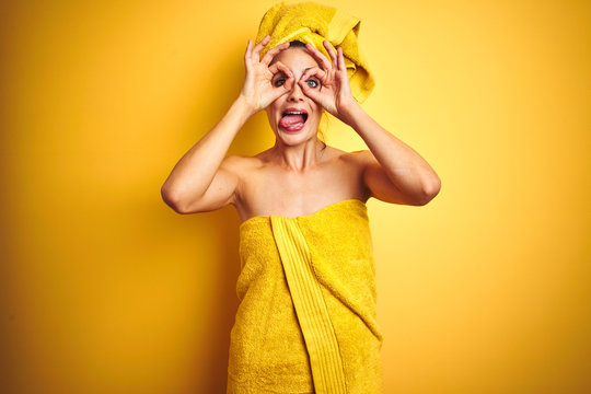 Beautiful woman wearing shower towel on body and head over yellow isolated background doing ok gesture like binoculars sticking tongue out, eyes looking through fingers. Crazy expression.