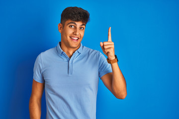 Fototapeta premium Young indian man wearing casual polo standing over isolated blue background pointing finger up with successful idea. Exited and happy. Number one.