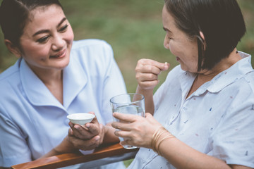 Asian nurse helping sick elderly woman to drink water with medical pill.