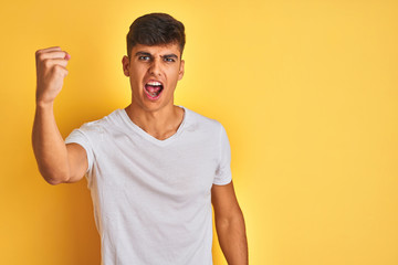 Young indian man wearing white t-shirt standing over isolated yellow background angry and mad...
