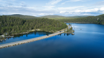 Aerial view of road between green forest and blue lake. Cars moving on road. Beautiful summer panorama.