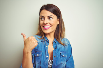 Young beautiful woman standing over isolated background smiling with happy face looking and pointing to the side with thumb up.