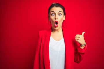Young beautiful business woman standing over red isolated background Surprised pointing with hand finger to the side, open mouth amazed expression.