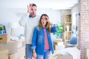 Young beautiful couple standing at new home around cardboard boxes doing ok gesture shocked with surprised face, eye looking through fingers. Unbelieving expression.