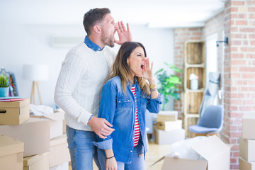 Young beautiful couple standing at new home around cardboard boxes shouting and screaming loud to side with hand on mouth. Communication concept.