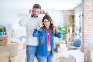 Young beautiful couple standing at new home around cardboard boxes covering ears with fingers with...