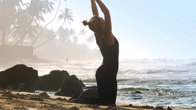 blonde girl with space buns does yoga backbend pose on warm morning ocean beach against palms in fog and rolling waves slow motion. Concept yoga sport, travel
