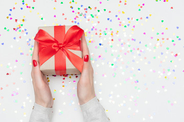 White desk with scattered tinsel, woman holds gift, woman's hands, gift box with red ribbon, close up