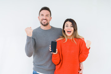 Young couple showing smartphone screen over isolated background screaming proud and celebrating victory and success very excited, cheering emotion