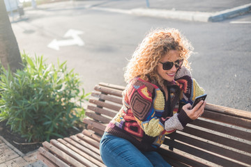 woman alone in the street sitting on a bench of the park using her phone and smiling at it - online lifestyle concept - curly blonde beautiful girl with technology device to stay connected 