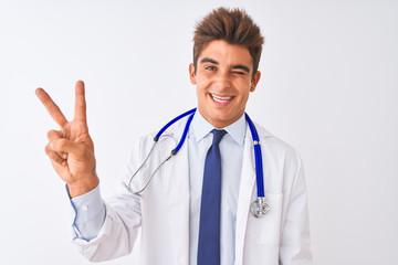 Young handsome doctor man wearing stethoscope over isolated white background smiling with happy face winking at the camera doing victory sign. Number two.