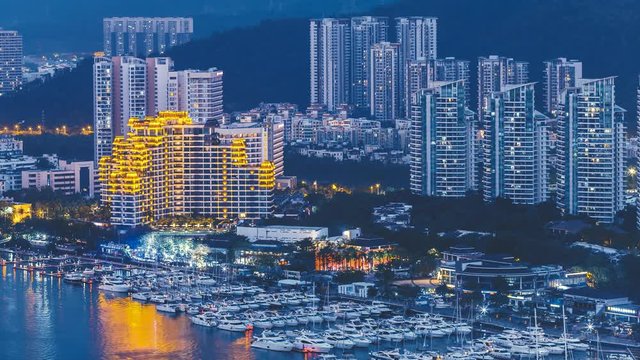 Haikou city with high modern buildings on Nandu River bank with mooring motorboats yachts getting illuminated in darkness time lapse. Concept travel transport, Hainan Province China. Cinematic 4K