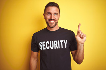 Young safeguard man wearing security uniform over yellow isolated background with a big smile on face, pointing with hand and finger to the side looking at the camera.