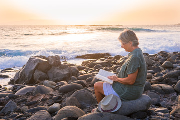 A senior woman with grey hair sitting alone on the beach of pebbles reading a book. 70 years old....
