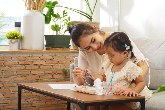 Beautiful Asian woman teaching her cute daughter painting ceramic models on the table using colorful watercolor in living room. Kid learning with imagination, happy family, lifestyle and love concept