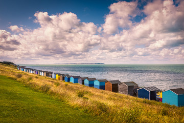 Row of beach huts under a white cloudscape along the coast in Tankerton, Whitstable, Kent. The grass slopes behind the huts are turning a golden yellow. Sheppey can be seen along the horizon.