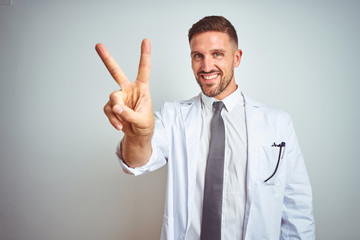 Young handsome doctor man wearing white profressional coat over isolated background smiling looking to the camera showing fingers doing victory sign. Number two.