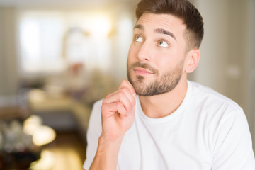 Obraz premium Young handsome man wearing casual white t-shirt at home with hand on chin thinking about question, pensive expression. Smiling with thoughtful face. Doubt concept.