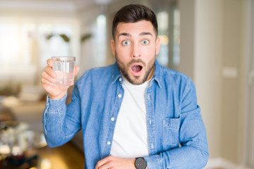 Young handsome man drinking a glass of water at home scared in shock with a surprise face, afraid and excited with fear expression