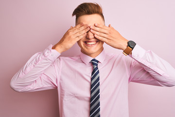 Young handsome businessman wearing shirt and tie standing over isolated pink background covering eyes with hands smiling cheerful and funny. Blind concept.