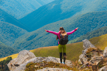 Young travelling girl with a backpack standing on the edge of a rock with raised hands admiring the beauty of landscape