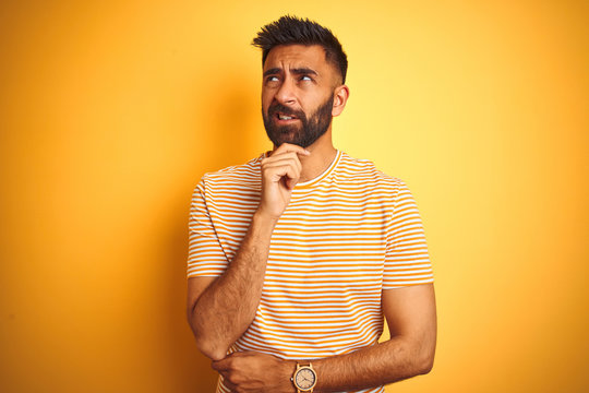 Young indian man wearing t-shirt standing over isolated yellow background Thinking worried about a question, concerned and nervous with hand on chin