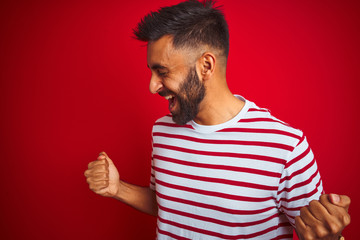 Young indian man wearing striped t-shirt standing over isolated red background very happy and excited doing winner gesture with arms raised, smiling and screaming for success. Celebration concept.