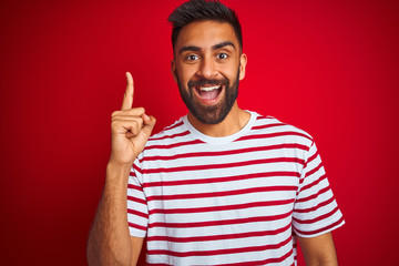Fototapeta premium Young indian man wearing striped t-shirt standing over isolated red background pointing finger up with successful idea. Exited and happy. Number one.
