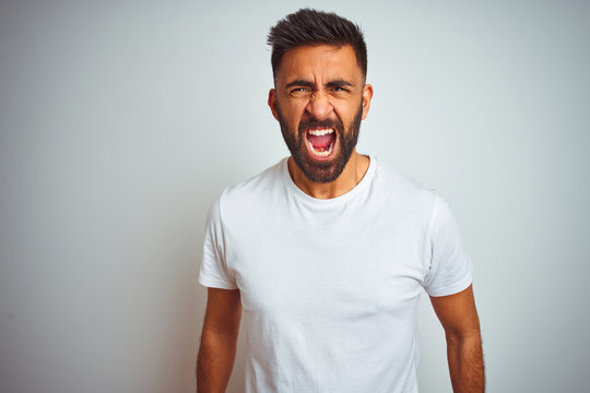 Young indian man wearing t-shirt standing over isolated white background angry and mad screaming frustrated and furious, shouting with anger. Rage and aggressive concept.