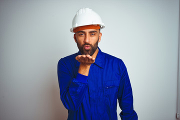 Handsome indian worker man wearing uniform and helmet over isolated white background looking at the camera blowing a kiss with hand on air being lovely and sexy. Love expression.