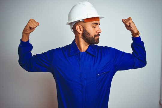 Handsome indian worker man wearing uniform and helmet over isolated white background showing arms muscles smiling proud. Fitness concept.