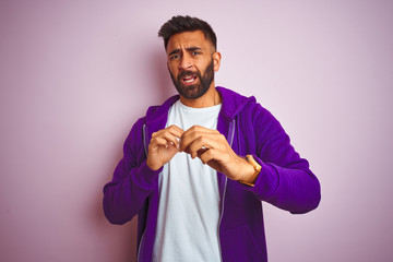 Young indian man wearing purple sweatshirt standing over isolated pink background disgusted expression, displeased and fearful doing disgust face because aversion reaction. With hands raised. 