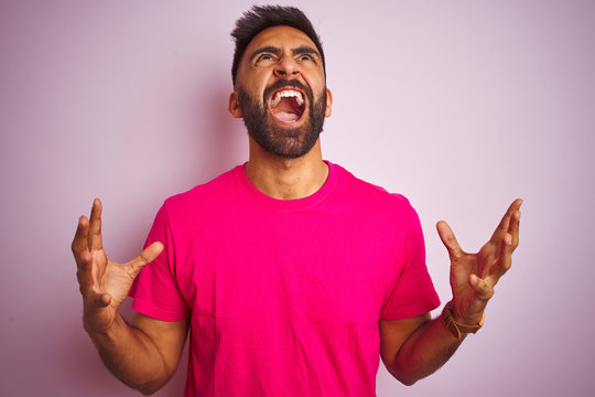 Young indian man wearing t-shirt standing over isolated pink background crazy and mad shouting and yelling with aggressive expression and arms raised. Frustration concept.