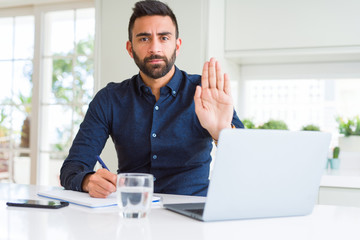 Handsome hispanic man working using computer and writing on a paper with open hand doing stop sign with serious and confident expression, defense gesture