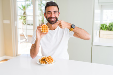 Handsome hispanic man eating sweet belgian waffle pastry very happy pointing with hand and finger