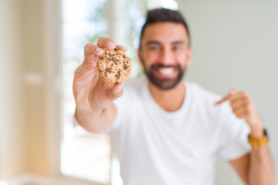 Handsome hispanic man eating chocolate chips cookies with surprise face pointing finger to himself