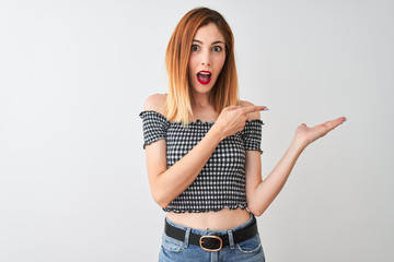 Beautiful redhead woman wearing casual t-shirt standing over isolated white background amazed and smiling to the camera while presenting with hand and pointing with finger.