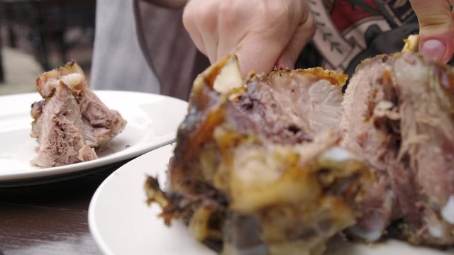 Man's hands with a knife and a fork cuts a pork knuckle and puts it on a plate. Nearby is a glass of beer.