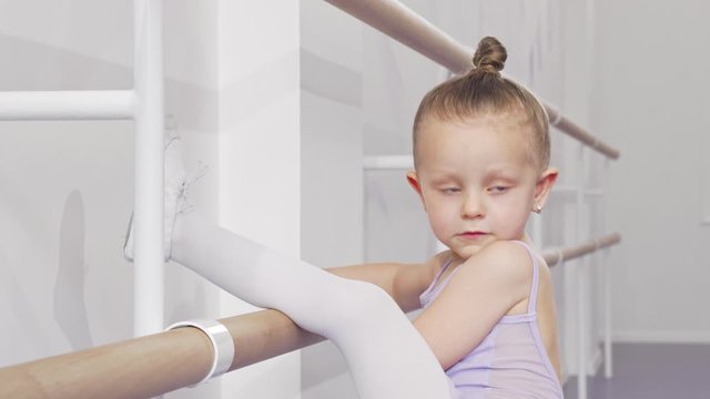 Adorable little ballerina girl putting her leg on ballet bar for stretching. Happy little girl in leotard exercising at ballet school, stretching her leg on barre. Flexibility, gymnastics concept