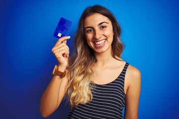 Young beautiful woman holding credit card over blue isolated background with a happy face standing and smiling with a confident smile showing teeth
