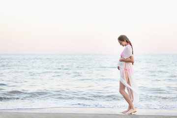 Young pregnant woman is standing on the seashore and hugging her belly. Enjoying the moment.