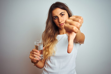 Young beautiful woman drinking a glass of water over white isolated background with angry face,...