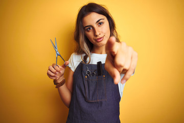 Young beautiful hairdresser woman holding scissors over yellow isolated background pointing with...
